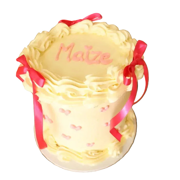 Decorative cake with pink ribbons and 'Maize' written on it, placed on a wooden table.