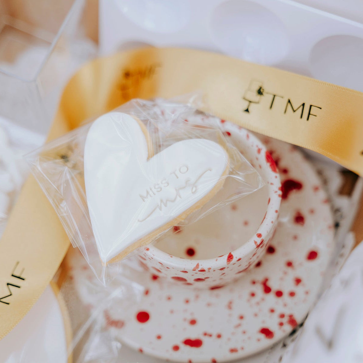 Heart-shaped cookie with white icing, wrapped in clear plastic, on a tea cup and saucer with red splashes, with a gold band featuring 'TMF'.