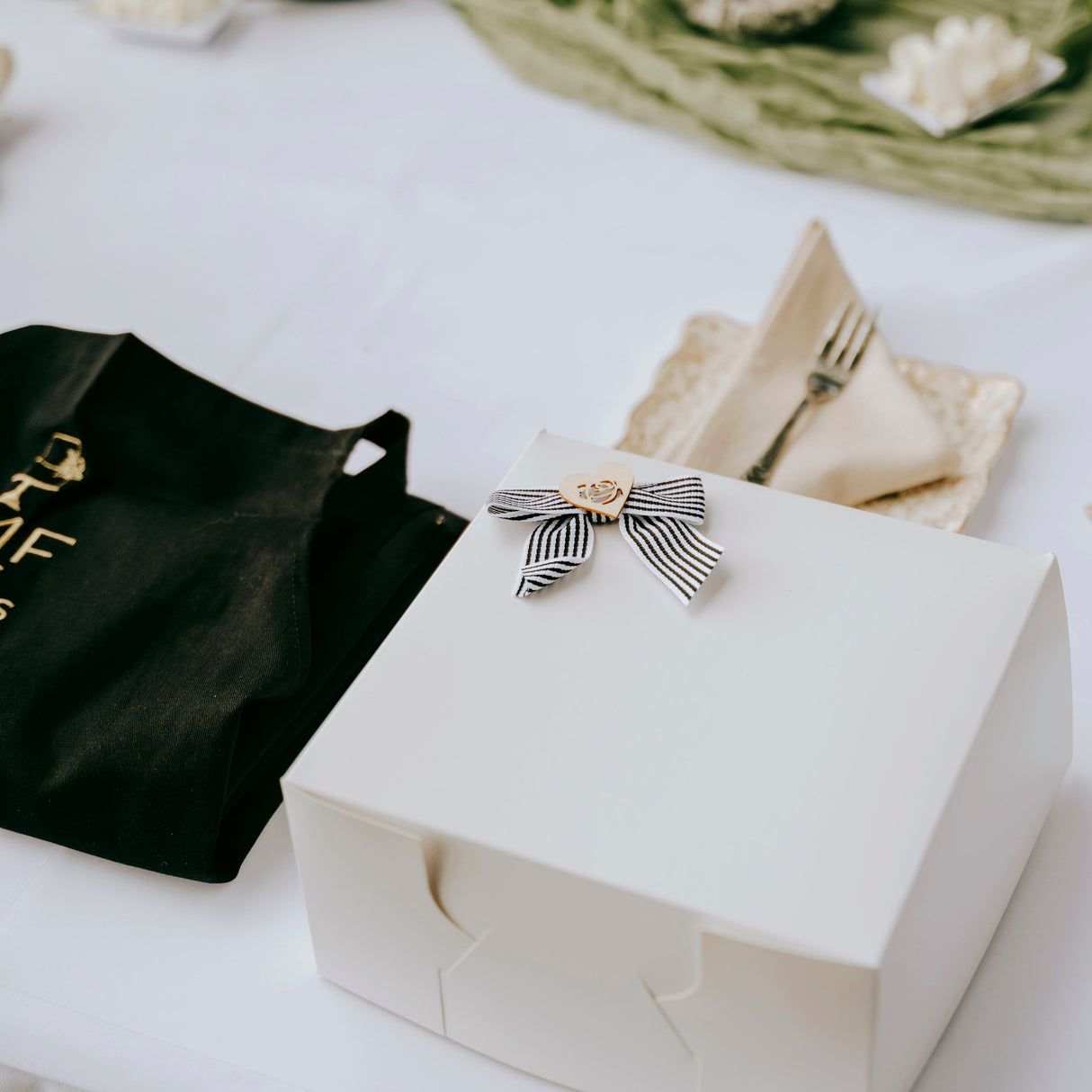 White box with a decorative bow on a table with a black TMF Apron and folded napkin.