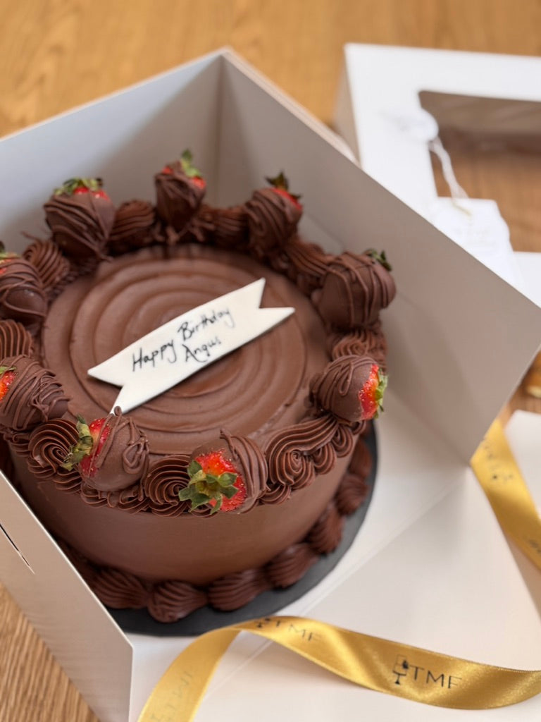 A belgian chocolate mud cake with chocolate ganache frosting and decorations, presented in a box with a 'Happy Birthday' message.