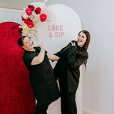 Two people posing in front of a 'Cake & Sip' sign with floral decorations.
