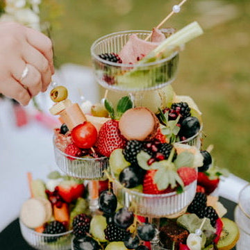 A three-tiered glass fruit platter filled with strawberries, blackberries, grapes, kiwi, and macarons, set outdoors on a white tablecloth with a person's hand reaching in to pick a fruit skewer.