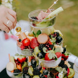 A three-tiered glass fruit platter filled with strawberries, blackberries, grapes, kiwi, and macarons, set outdoors on a white tablecloth with a person's hand reaching in to pick a fruit skewer.