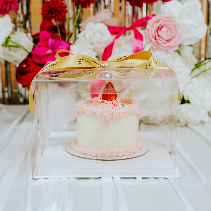 Small cake with a strawberry on top, placed under a clear acrylic cover with a gold bow, surrounded by flowers.
