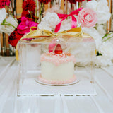 Small cake with a strawberry on top, placed under a clear acrylic cover with a gold bow, surrounded by flowers.