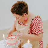 Person in a checkered shirt and apron preparing a small cake on a table with flowers and candles.