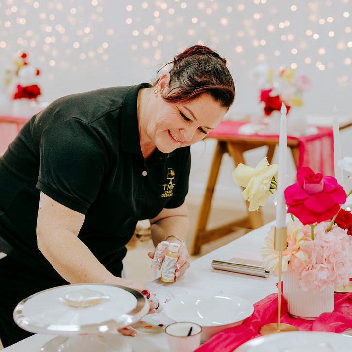 Person setting a table with floral arrangements and candles in a softly lit room.
