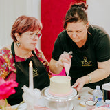 Two women decorating a cake at a table with flowers and a red heart-shaped decoration in the background.
