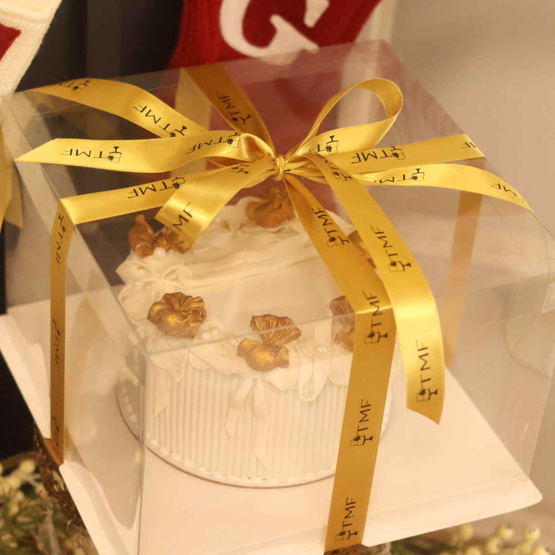 White cake with gold decorations in a clear box with a gold ribbon, blurred background