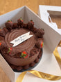 A belgian chocolate mud cake with chocolate ganache frosting and decorations, presented in a box with a 'Happy Birthday' message.